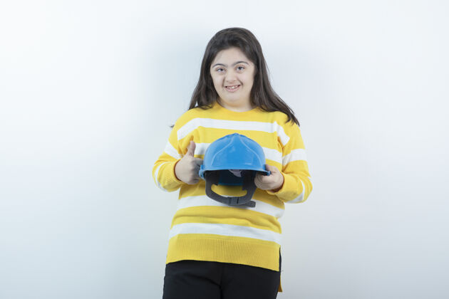 残疾穿条纹毛衣的可爱女孩 白墙上戴着蓝色的安全帽戴艺术家头盔