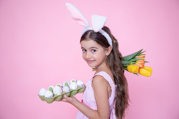 托盘有复活节兔子耳朵的小女孩手里拿着一束郁金香和一盘鸡蛋放在粉红色的墙上女孩春天兔子