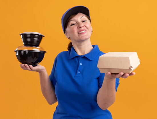 快乐身穿蓝色制服 头戴鸭舌帽的中年女送货员站在橘色的墙上 手里拿着食品包 开心而积极地看着前面食物盒子女人