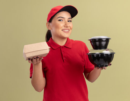 帽子身着红色制服 头戴鸭舌帽 面带微笑地站在绿色墙壁上 拿着食品包的快乐的年轻送货员望着前方女人快乐制服