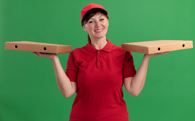 盒子身穿红色制服 头戴鸭舌帽的中年送货妇女站在绿色的墙上 面带微笑地看着前面的比萨饼盒女人抱着开心