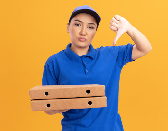 制服身穿蓝色制服 头戴鸭舌帽的年轻女送货员拿着比萨饼盒 不高兴地朝前竖起大拇指站在橙色的墙上盒子放下展示