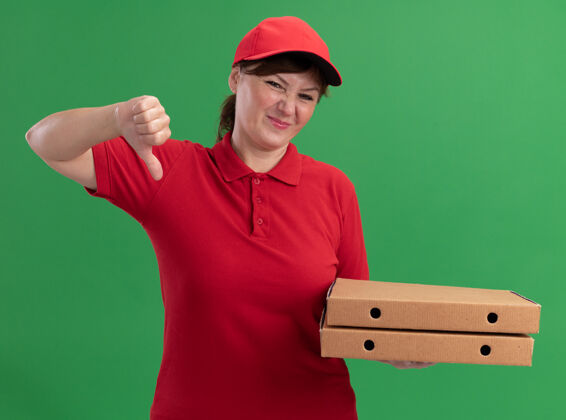 拇指身穿红色制服 头戴鸭舌帽的中年女送货员拿着披萨盒 困惑地看着前面 竖起大拇指站在绿色的墙上年龄中间表演
