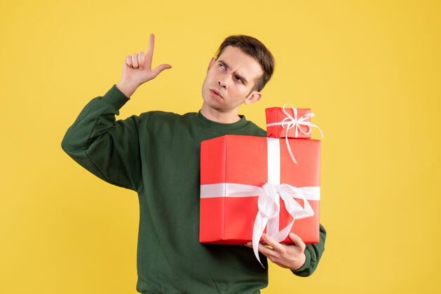 人物正面图：年轻人拿着圣诞礼物指着天花板站在黄色背景上成人黄色快乐