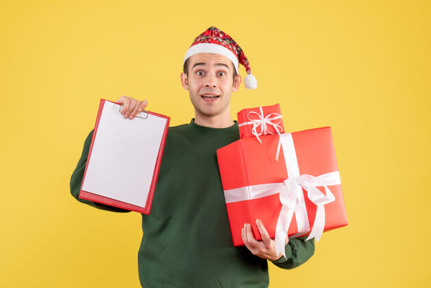 视图正面图：戴着圣诞帽的年轻人拿着礼物和剪贴板站在黄色背景上男人前面礼物