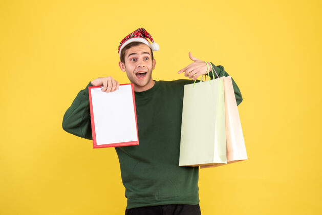 前面正面图：戴着圣诞帽的年轻人拿着购物袋和剪贴板站在黄色背景上站男人微笑