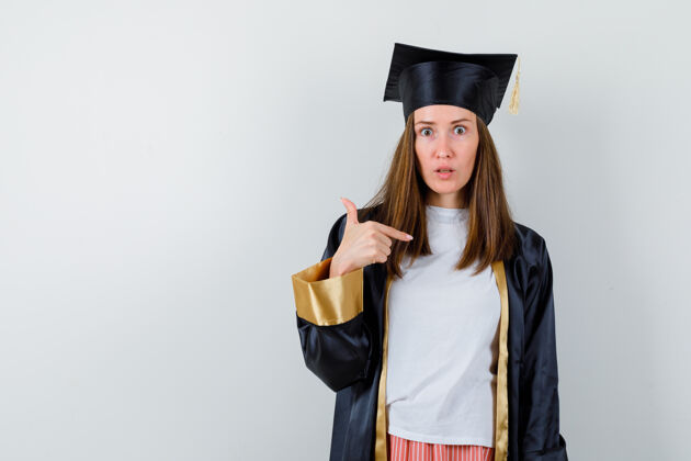 长袍毕业妇女的肖像指着自己在休闲服 制服 看着困惑的前视图成人教育休闲