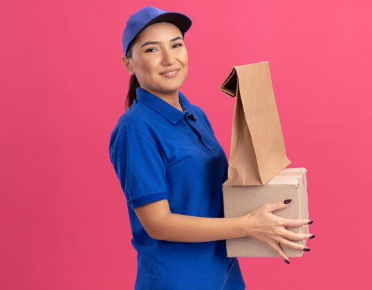 女人身着蓝色制服 头戴鸭舌帽 拿着纸包和纸板箱的快乐年轻女送货员站在粉色的墙上 微笑着自信地看着前方纸站立纸板