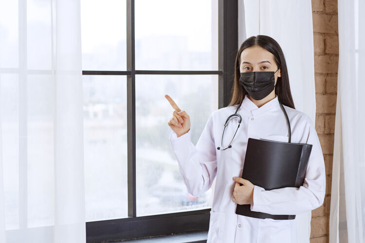 护理带着听诊器和黑色面罩的医生站在窗边 拿着病人的黑色病历夹 指着某处健康治疗检查表