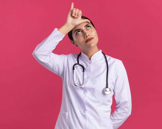 听诊器年轻的女医生 穿着医用外套 手持听诊器 脸上带着恼怒的表情抬起头来 站在粉色的墙上做着失败者的手势医生手势制造