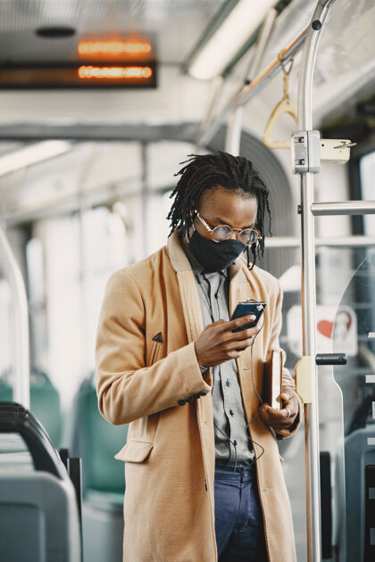人非裔美国人坐在城市巴士上穿棕色外套的家伙电晕病毒概念手机男子成人