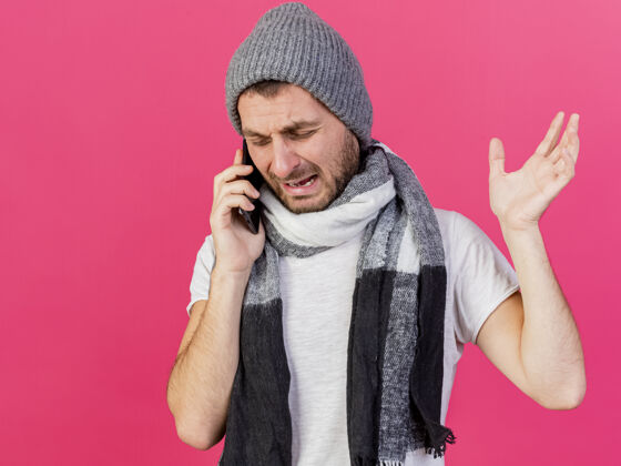 说话带着围巾戴着冬天帽子的哭泣的生病的年轻人在电话里讲话 举起孤立在粉色背景上的手疾病围巾帽子