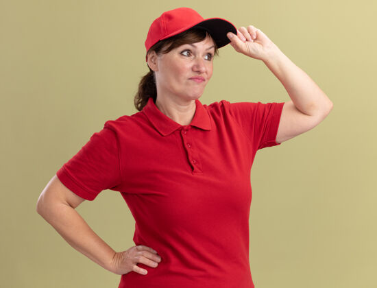 中间身穿红色制服 戴着帽子的中年女送货员站在绿色的墙上 困惑地 不高兴地碰了碰帽子分娩女人年龄