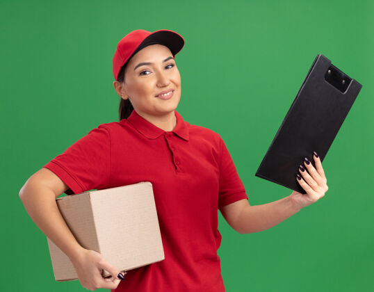 帽子身穿红色制服 头戴鸭舌帽的年轻女送货员拿着带剪贴板的纸板箱望着前方 满脸笑容自信地站在绿色的墙上微笑制服交货