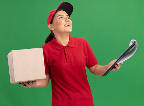 帽子身穿红色制服 头戴鸭舌帽的年轻送货员站在绿色的墙上 面带微笑地抬起头来 手里拿着一个纸板箱 上面夹着剪贴板女人纸板交货