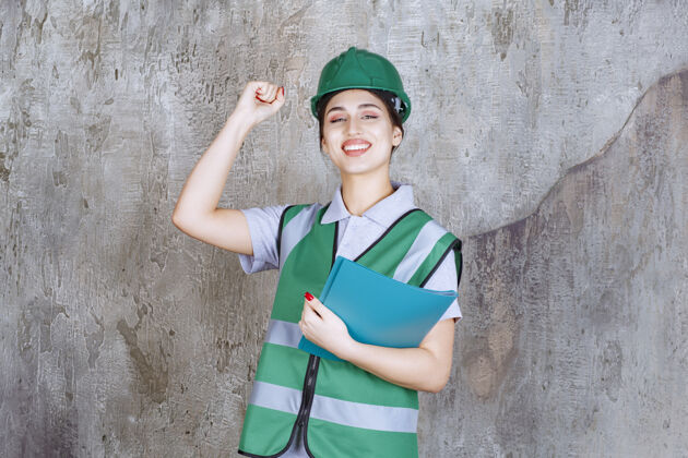 建筑身穿绿色制服 头戴安全帽的女工程师手持蓝色项目文件夹 并显示积极的手势满意姿势快乐