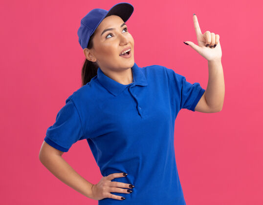 欢呼身穿蓝色制服 头戴帽子的年轻送货员抬起头来 用食指指着站在粉色墙上的某个微笑着的东西制服帽子什么