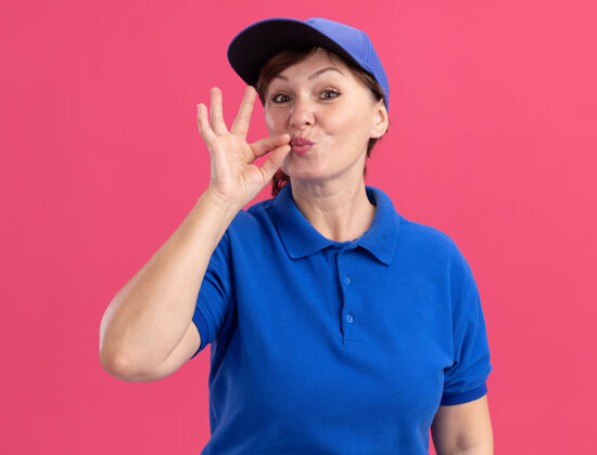 关闭身穿蓝色制服 头戴鸭舌帽的中年女送货员沉默地做着一个手势 像是站在粉色墙壁上用拉链合上嘴巴沉默中间帽子