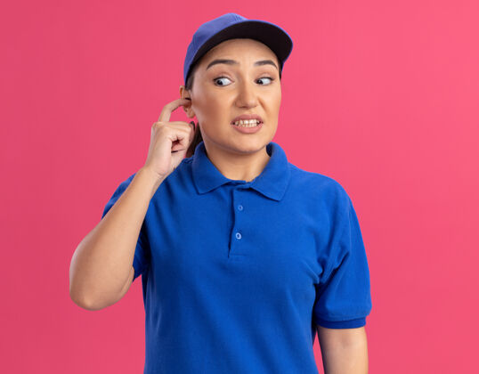 表情穿着蓝色制服 戴着帽子的年轻女送货员站在粉红色的墙上 困惑地用手指合上耳朵 脸上露出恼怒的表情帽子站立烦恼