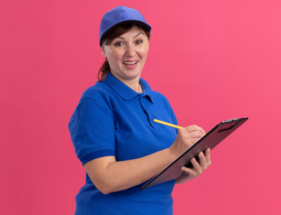 写字身穿蓝色制服 头戴鸭舌帽的中年女送货员拿着写字板和铅笔 站在粉红色的墙边 愉快地看着前面的幸福和积极的微笑欢呼制服正面