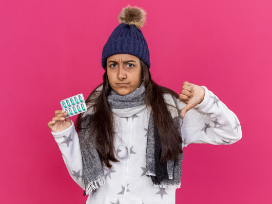 药片未被释放的生病的年轻女孩戴着冬天的帽子和围巾拿着药片 显示拇指向下孤立的粉红色背景穿疾病下来