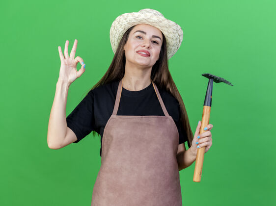 制服微笑着美丽的园丁女孩穿着制服 戴着园艺帽 拿着耙子 在绿色的背景上表现出很好的姿态帽子花园手势