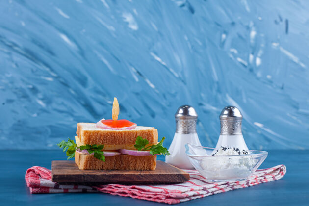 欧芹三明治放在一块木板上 旁边放着盐和一碗奶酪 放在茶巾上 放在蓝色的桌子上奶酪美味芝麻
