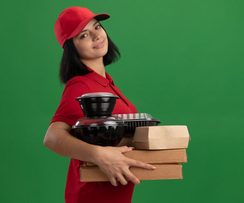 送货身穿红色制服 头戴鸭舌帽 手持比萨饼盒和食品包的年轻送货女孩站在绿色的墙上 脸上洋溢着幸福的笑容女孩微笑制服