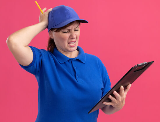 中间身穿蓝色制服 头戴帽子的中年送货妇女手举铅笔 手放在头上 失望地站在粉红色的墙上剪贴板头铅笔