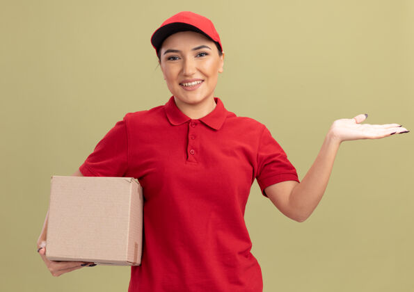帽子身穿红色制服 头戴鸭舌帽的年轻送货员拿着纸板箱 微笑着自信地看着前面的复印空间 她的手臂站在绿色的墙上手臂介绍信心