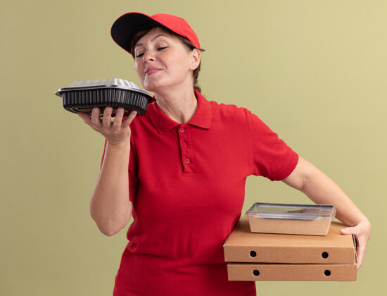 香味身着红色制服 头戴鸭舌帽的中年女送货员手持披萨盒和食品包装 站在绿色的墙边 愉快而积极地吸入怡人的香气送货帽子愉快