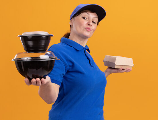 微笑身着蓝色制服 头戴鸭舌帽的中年女送货员站在橙色的墙上 脸上露出自信的微笑盒子食物女人