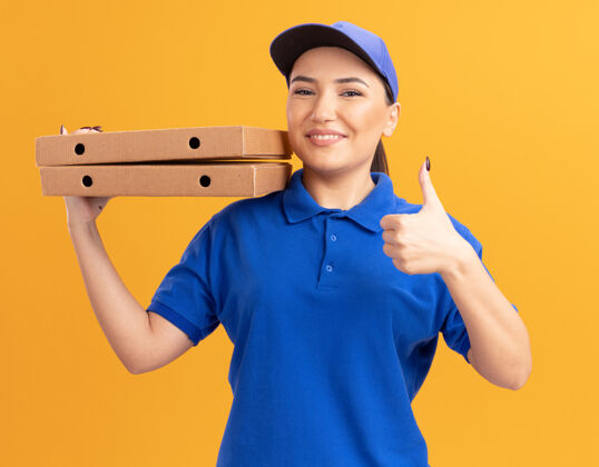 送货快乐的年轻送货员身穿蓝色制服 戴着帽子 拿着披萨盒 微笑自信地站在橙色的墙上竖起大拇指 看着前面拇指抱着制服