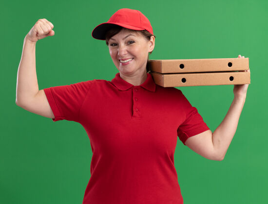 站着快乐的中年送货员 穿着红色制服 戴着帽子 手里拿着披萨盒 举起拳头看着前面 微笑着 自信地站在绿色的墙上披萨抱着自信