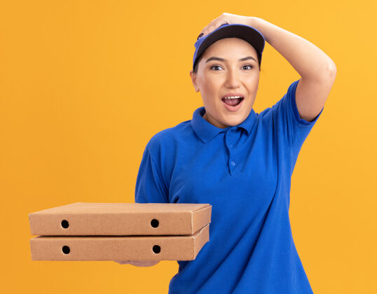 站着身穿蓝色制服 戴着帽子 拿着披萨盒的年轻女送货员站在橙色的墙上 惊讶地看着前面帽子制服年轻人