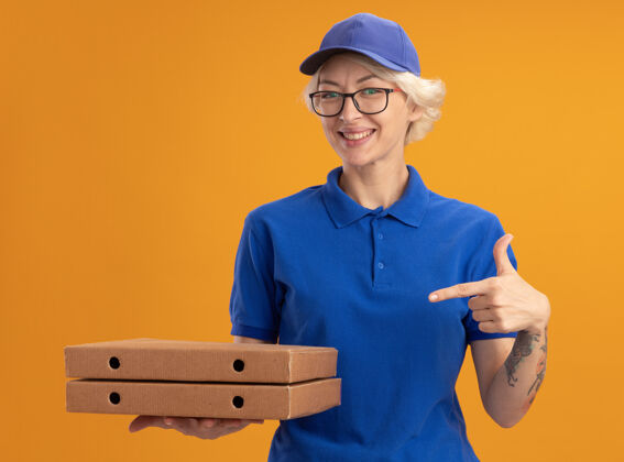 手指身穿蓝色制服 戴着眼镜 拿着比萨饼盒的年轻送货员用食指指着他们 在橙色的墙上欢快地微笑着年轻人欢呼戴着