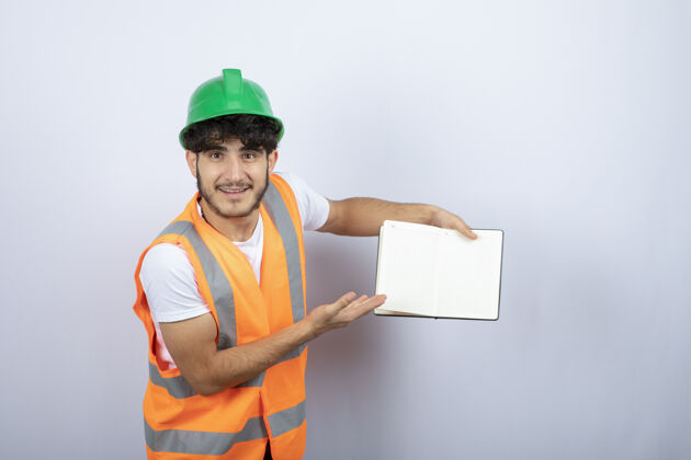 帽子戴绿色安全帽的年轻男工程师在白色背景上展示笔记高质量照片建筑工程师制造