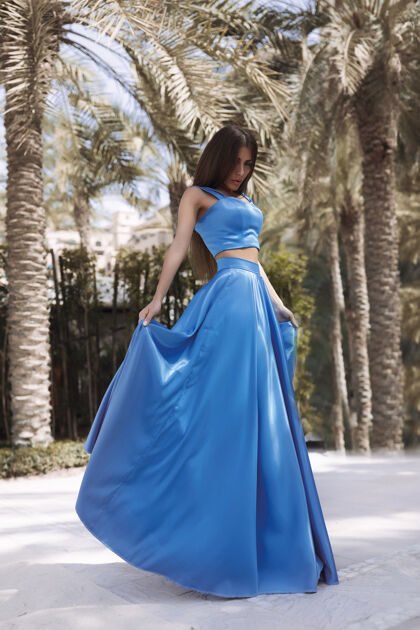 时尚的外观美丽性感的黑发女郎穿着蓝色的长裙站在泳池边 长发 完美的身材和漂亮的脸蛋 化妆 在棕榈树下身材连衣裙鸡尾酒礼服