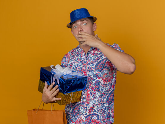 盖害怕的年轻人戴着蓝色帽子拿着礼品盒包嘴手孤立在橙色背景害怕帽子手