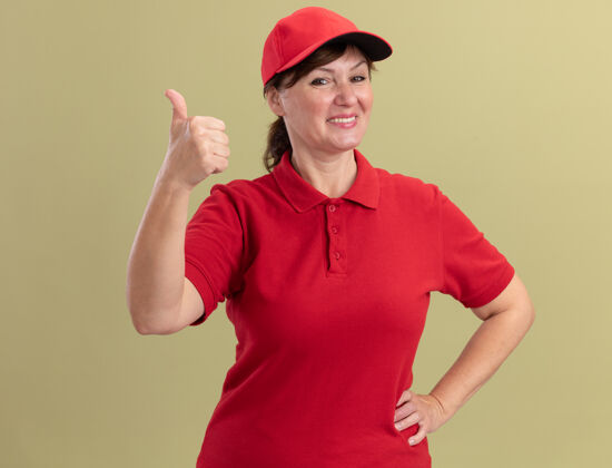 站着身穿红色制服 头戴帽子的中年女送货员站在绿色的墙上 面带微笑 高高兴兴地竖起大拇指送货欢呼女人