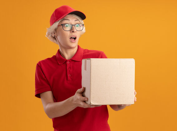 站着快乐的年轻送货女 穿着红色制服 戴着帽子 戴着眼镜 拿着纸板箱 站在橘色的墙上惊讶地看着它制服眼镜穿上