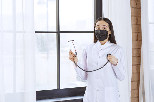 检查戴着黑色口罩的医生站在窗边用听诊器检查心跳感染妇女诊所