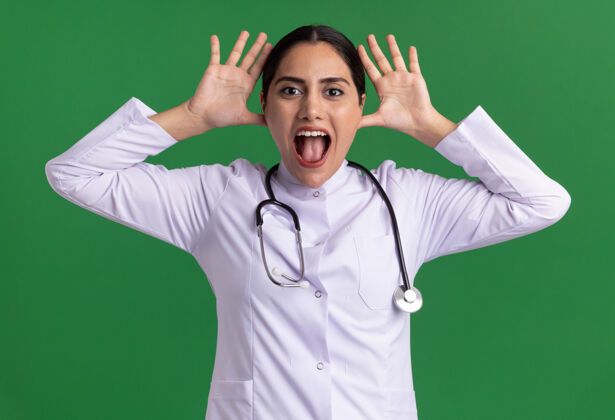 舌头有趣的年轻女医生 穿着医用外套 手持听诊器 站在绿色的墙壁上 看着前方 做鬼脸 伸出舌头 双手靠近头部站着鬼脸搞笑