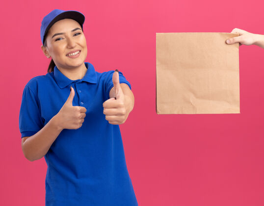 展示身着蓝色制服 戴着帽子的年轻送货员站在粉红色的墙上 友好地竖起大拇指 一边接受纸袋站立递送拇指