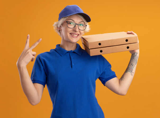 年轻人穿着蓝色制服 戴着帽子 戴着眼镜 拿着披萨盒 微笑着在橙色的墙上画着v字标志的快乐的年轻女送货员眼镜拿着制服