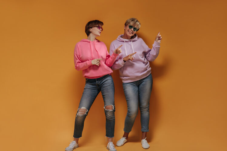中等两位短发女士的全幅照片 戴着明亮的太阳镜 淡紫色和粉色的连帽衫 牛仔裤 在橙色的背景上展示文字的位置女老年人衬衫