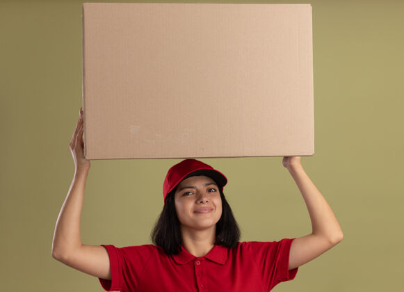轻身穿红色制服 头戴鸭舌帽 头上抱着大纸箱 友好微笑地站在轻质墙上的年轻送货女孩朋友大举行
