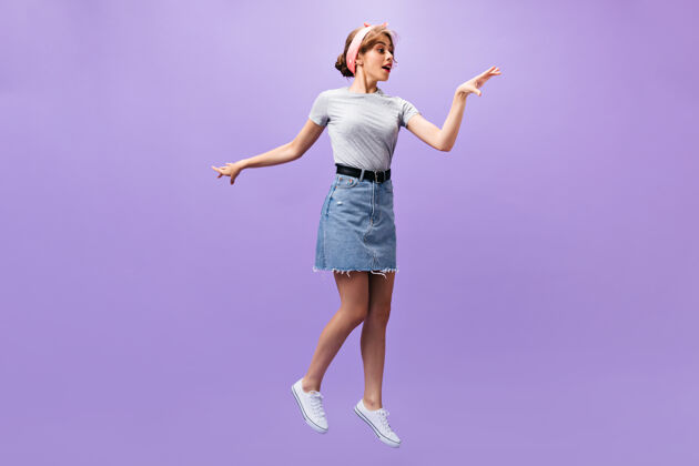 衬衫穿着牛仔裙的迷人女人在紫色背景上跳跃穿着粉色头带和灰色衬衫的时髦漂亮女孩摆姿势一件紫色女孩