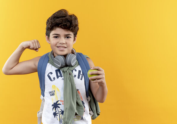 戴着高兴的小男孩戴着书包和耳机拿着苹果和显示是的手势隔离在黄色背景与复制空间拿着展示苹果