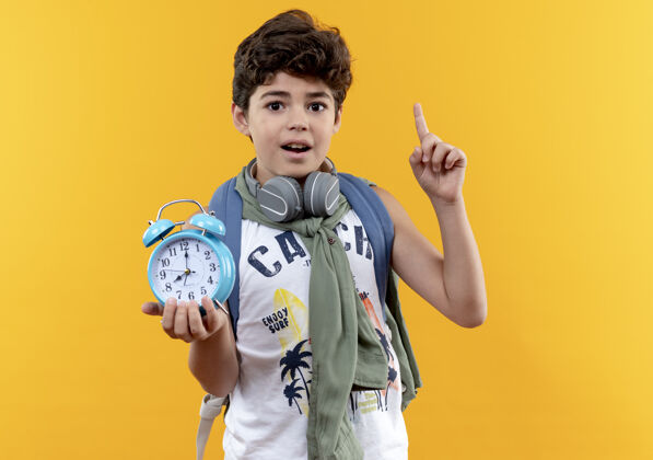 耳机给人印象深刻的小男生背着书包 戴着耳机 拿着闹钟 指着黄色背景上的孤立起来闹钟点抱